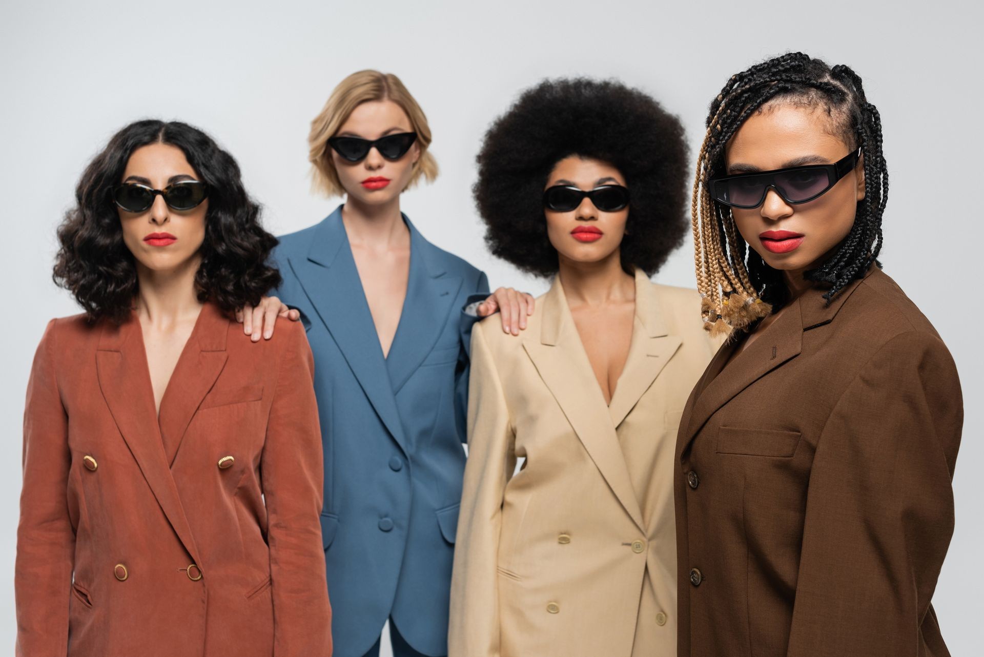 Four women wearing sunglasses and stylish blazers in different colors stand confidently against a neutral background.