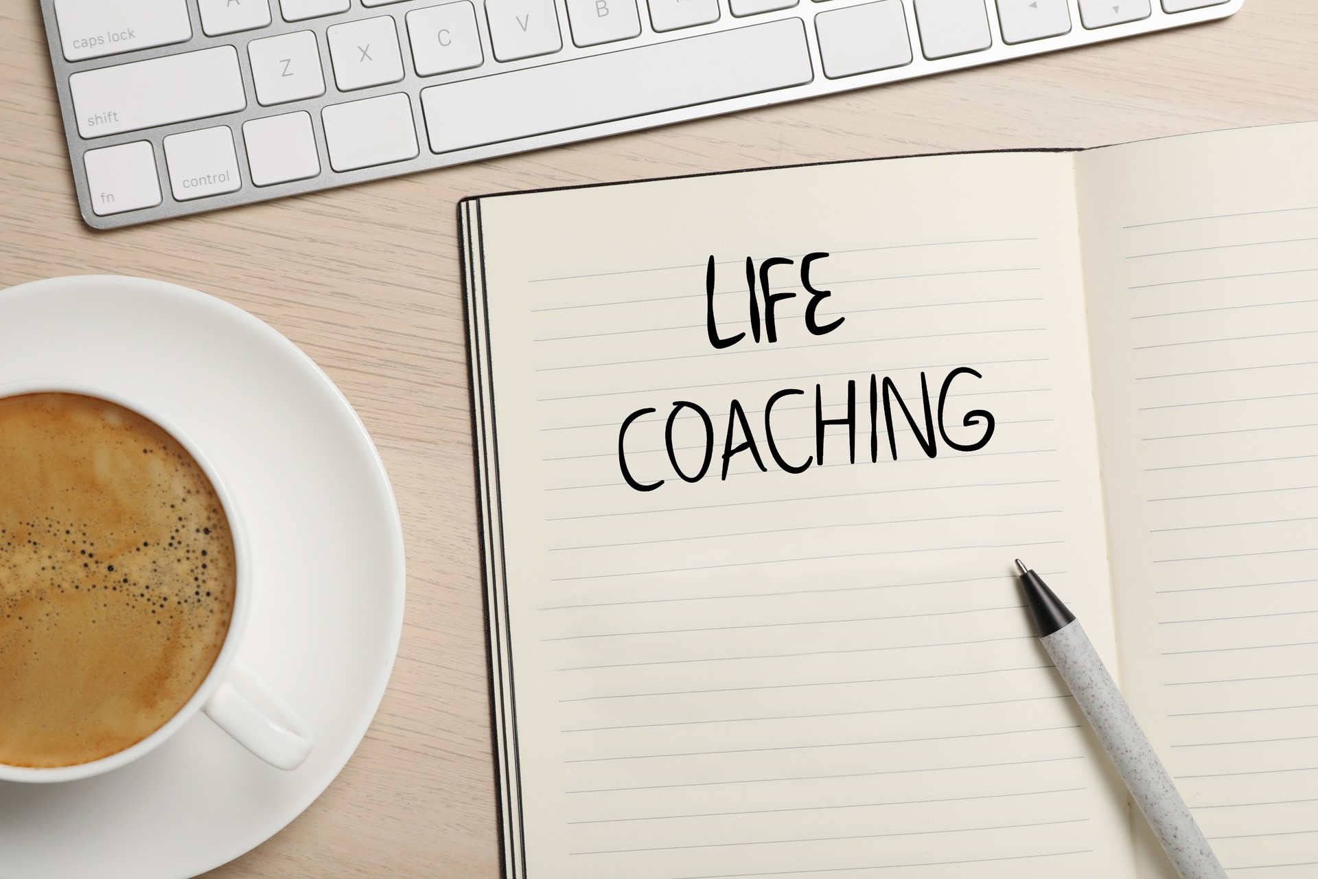 Phrase Life Coach written in notebook, pen, cup of coffee and keyboard on wooden table, flat lay